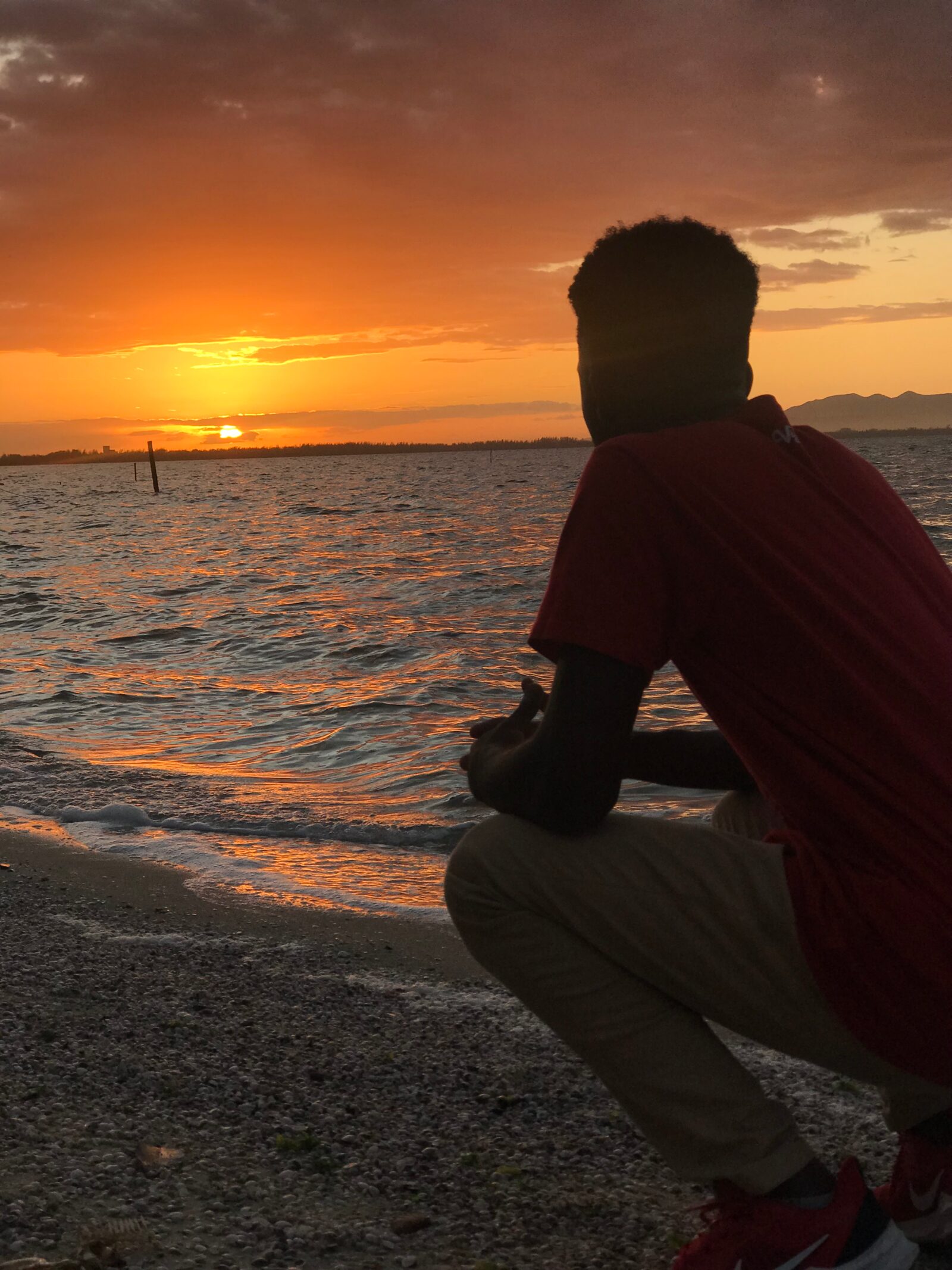 Moises Rosa agachado na beira do mar, de costas, observando o pôr do sol.